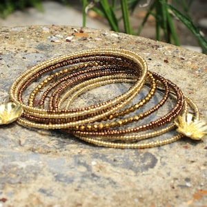 Könnte beinhalten: Ein goldfarbenes und braunes Perlenarmband mit zwei goldenen Blumenanhängern. Das Armband besteht aus mehreren Perlensträngen, die übereinander gelegt sind.