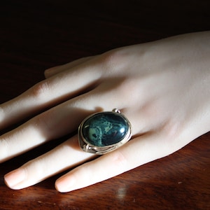 May include: A silver ring with an oval, dark green stone. The ring is set on a hand with a dark wooden background. The stone has a marbled appearance with lighter green and black swirls. The ring's setting has a decorative, ornate design.