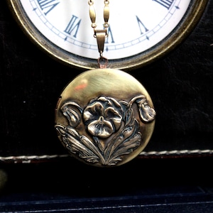 May include: A vintage-style brass locket necklace with a floral design. The round pendant features a detailed embossed flower and leaf pattern. The necklace hangs from a delicate chain, showcasing a classic and timeless jewelry piece.