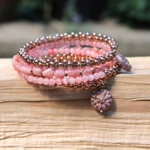 Könnte beinhalten: Ein mehrreihiges Armband mit rosa und kupferfarbenen Perlen. Das Armband ist um ein Stück Holz gewickelt. Das Armband hat zwei dekorative Anhänger.