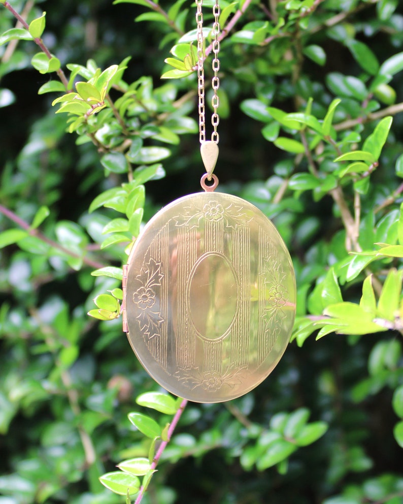 May include: A gold oval locket with an engraved design and a chain. The locket is hanging from a branch of a green bush.