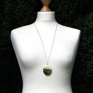 May include: A gold necklace with a large, oval pendant. The pendant has a textured surface and is hanging on a white mannequin.