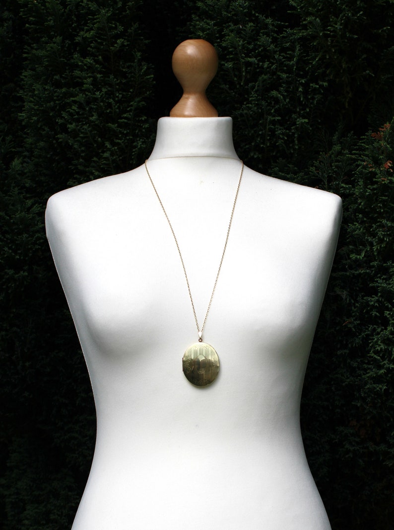 May include: A gold necklace with a large, oval pendant. The pendant has a textured surface and is hanging on a white mannequin.