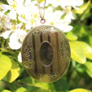May include: A gold locket necklace with a detailed floral design and a striped pattern on the front. The locket is hanging from a delicate gold chain.