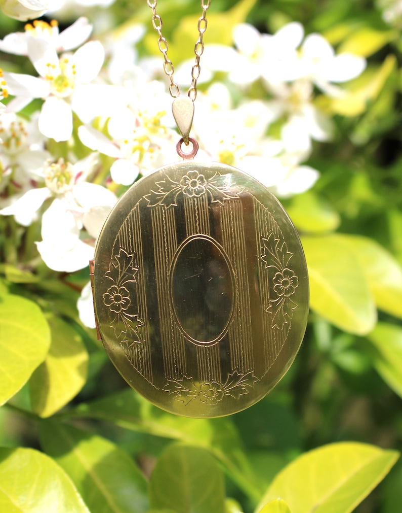 May include: A gold locket necklace with a detailed floral design and a striped pattern on the front. The locket is hanging from a delicate gold chain.