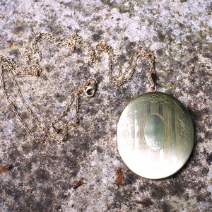 May include: A gold locket necklace with a detailed engraved design. The locket is oval shaped and has a hinged opening. The chain is delicate and has a lobster clasp.