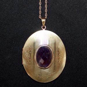 Puede incluir: Colgante de medallón ovalado dorado con una piedra preciosa púrpura facetada en el centro. El medallón tiene detalles de líneas verticales y grabados florales. Cuelga de una delicada cadena dorada.