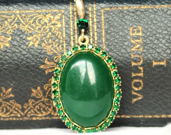 Puede incluir: Un colgante de oro con una piedra verde ovalada grande rodeada por un borde de piedras verdes más pequeñas.