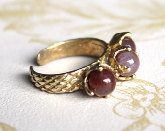 May include: A gold ring with three red and purple stones set in the band. The ring has a braided design.
