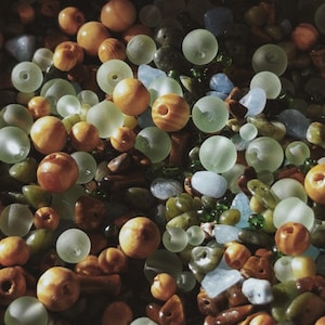May include: An overhead shot of a collection of beads and stones in various shapes and sizes. The beads are primarily round, in shades of amber, and frosted green. The stones are a mix of blues, greens, and browns, creating a textured, natural aesthetic.