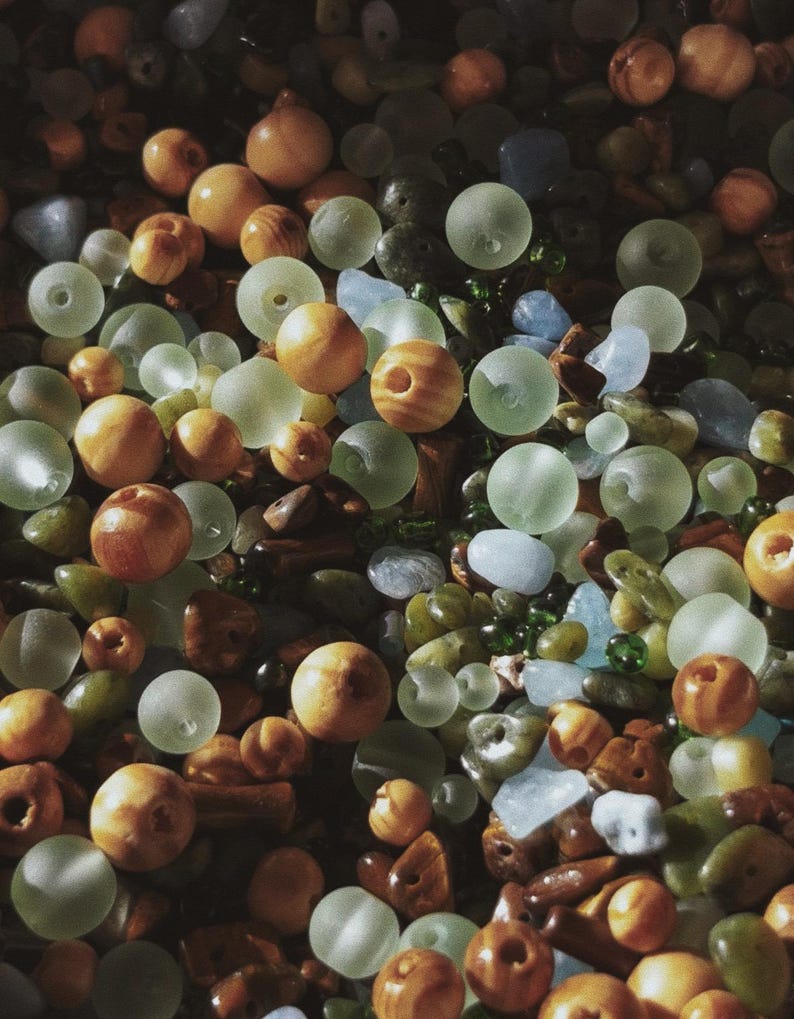 May include: An overhead shot of a collection of beads and stones in various shapes and sizes. The beads are primarily round, in shades of amber, and frosted green. The stones are a mix of blues, greens, and browns, creating a textured, natural aesthetic.
