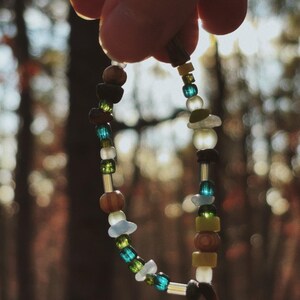 May include: A beaded bracelet with a variety of colorful beads, including blue, green, brown, and white. The bracelet is held up against a blurred background of trees. The beads are of different shapes and sizes, strung together on a thin cord.