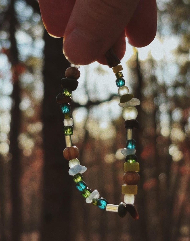 May include: A beaded bracelet with a variety of colorful beads, including blue, green, brown, and white. The bracelet is held up against a blurred background of trees. The beads are of different shapes and sizes, strung together on a thin cord.
