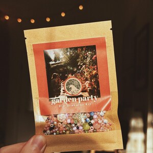 May include: A brown paper packet labeled "Garden Party DIY Bracelet Kit" with a red border and a photo of a garden. The packet contains a variety of colorful beads in shades of pink, green, and clear.