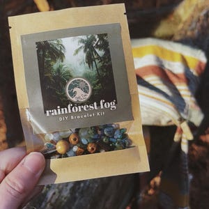 May include: A DIY bracelet kit in a clear plastic bag. The kit is labeled "rainforest fog" and includes beads in shades of green, blue, and brown. The bag has a picture of a rainforest scene. The kit is held by a person.