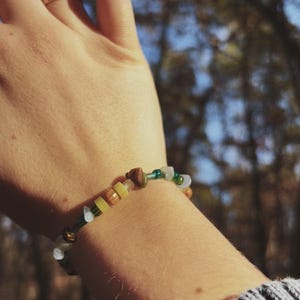 May include: A beaded bracelet with a mix of colors, including green, yellow, brown, and white beads. The bracelet is worn on a wrist, with a gold ring on a finger. The background is blurred, with trees and sunlight visible.