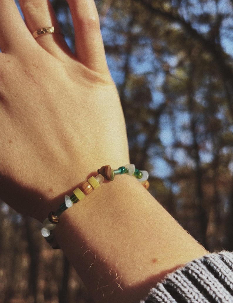 May include: A beaded bracelet with a mix of colors, including green, yellow, brown, and white beads. The bracelet is worn on a wrist, with a gold ring on a finger. The background is blurred, with trees and sunlight visible.
