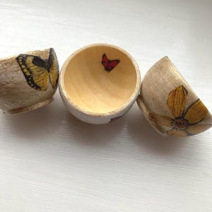 May include: Three small, wooden bowls with a light beige exterior and natural wood interior. Each bowl features a different nature-themed design: a yellow butterfly, a red butterfly, and a yellow flower. The bowls are arranged on a white surface.