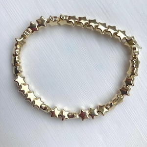 May include: A gold-coloured bracelet composed of star-shaped beads. The beads are strung together to form a complete circle. The stars are evenly spaced and have a shiny finish. The bracelet is displayed on a white surface.