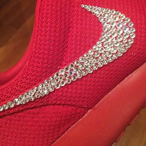May include: A red athletic shoe with a sparkling, crystal-studded design forming a swoosh logo on the side. The shoe features a textured fabric upper and a solid red sole, showcasing a blend of sporty and glamorous elements.