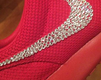May include: A red trainer with a sparkling, crystal-studded design forming a swoosh logo on the side. The shoe has a textured fabric upper and a solid red sole, blending sporty and glamorous elements. The design is eye-catching.