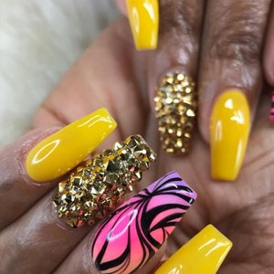 May include: Close-up of a hand with long, yellow nails. Some nails have gold rhinestones, and one nail has a pink and black swirl design.