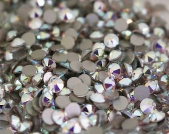 May include: A close-up shot of a collection of iridescent rhinestones. Each rhinestone has a flat, silver base and a clear, faceted top, creating a rainbow effect. These small decorative gems are used for crafts and embellishments.