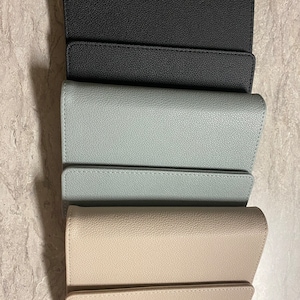 Pode incluir: Tr&ecirc;s carteiras de couro em preto, azul claro e bege. As carteiras est&atilde;o fechadas e empilhadas uma em cima da outra.