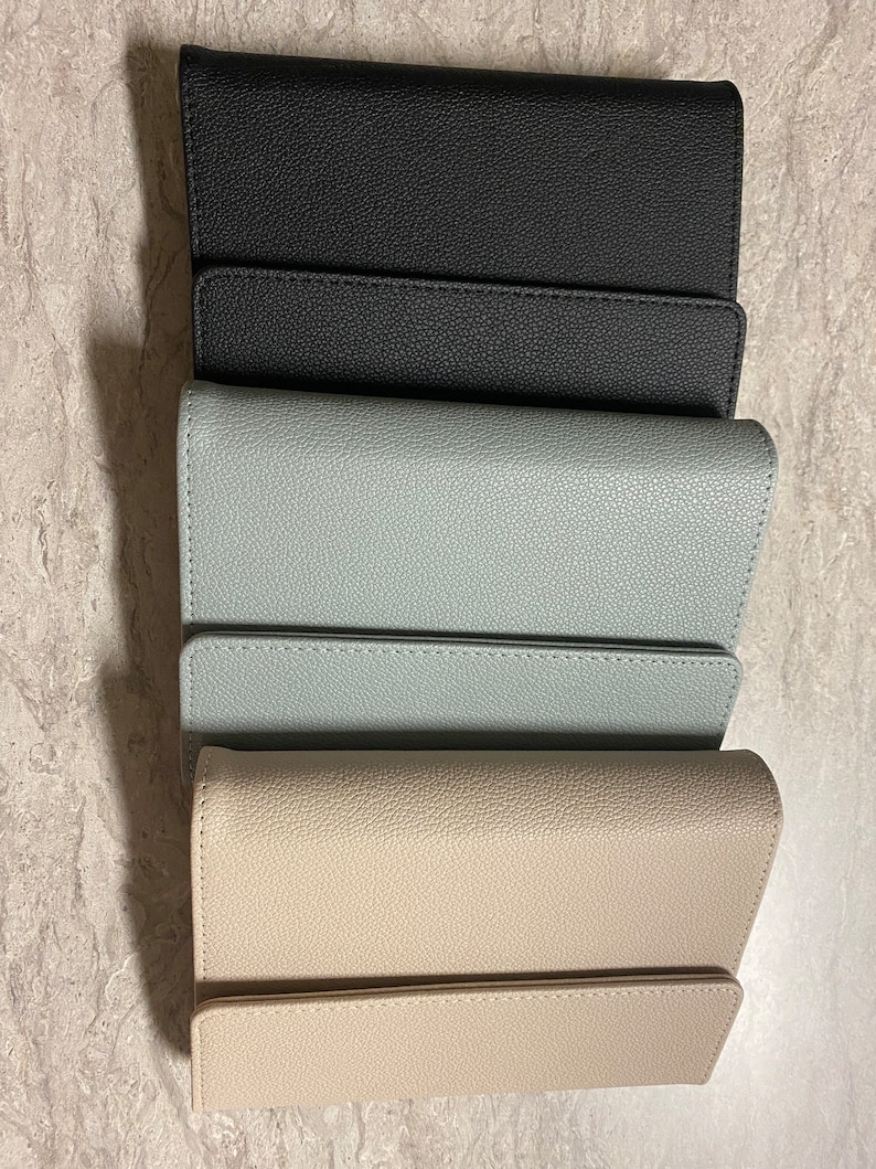 Pode incluir: Tr&ecirc;s carteiras de couro em preto, azul claro e bege. As carteiras est&atilde;o fechadas e empilhadas uma em cima da outra.