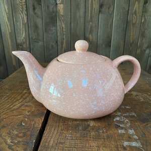May include: A pink speckled teapot with a white lid and a white handle. The teapot is sitting on a wooden surface.