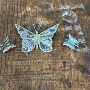 May include: Three silver butterfly brooches with iridescent abalone shell wings. The largest butterfly is in the center, with two smaller butterflies on either side.