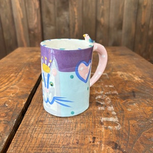 May include: A purple and blue ceramic mug with a cat face and a pink heart. The mug has a pink handle and a small bird perched on the rim.