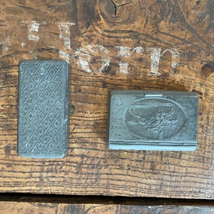 May include: Two vintage metal boxes. The box on the left has a diamond pattern on the front. The box on the right has a raised image of a person riding a horse and the word "BENG" on the front.