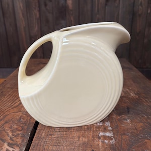 May include: A cream-colored ceramic pitcher with a rounded shape and a wide, flared spout. The pitcher has a smooth, glossy finish and a series of concentric circles around the body.