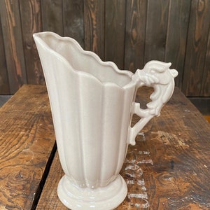 May include: A pale gray ceramic pitcher with a fluted body and a decorative handle shaped like a dragon's head. The pitcher is sitting on a wooden surface.
