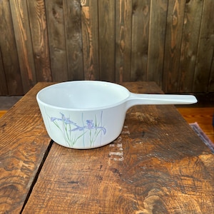 May include: White ceramic saucepan with a floral design featuring blue irises and green leaves. The saucepan has a white handle.