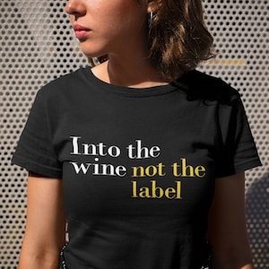 以下が含まれることがあります： 白い文字と金色の文字で「Into the wine not the label」と書かれた黒いTシャツです。