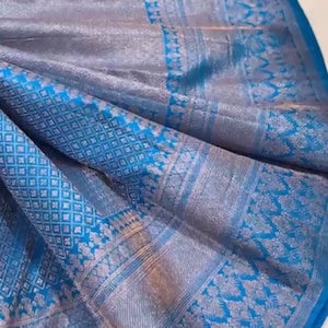 May include: A close-up of a blue and silver silk sari with intricate woven patterns. The fabric has a shimmering quality, with detailed floral and geometric designs. The sari is folded, showcasing its texture and color variations.
