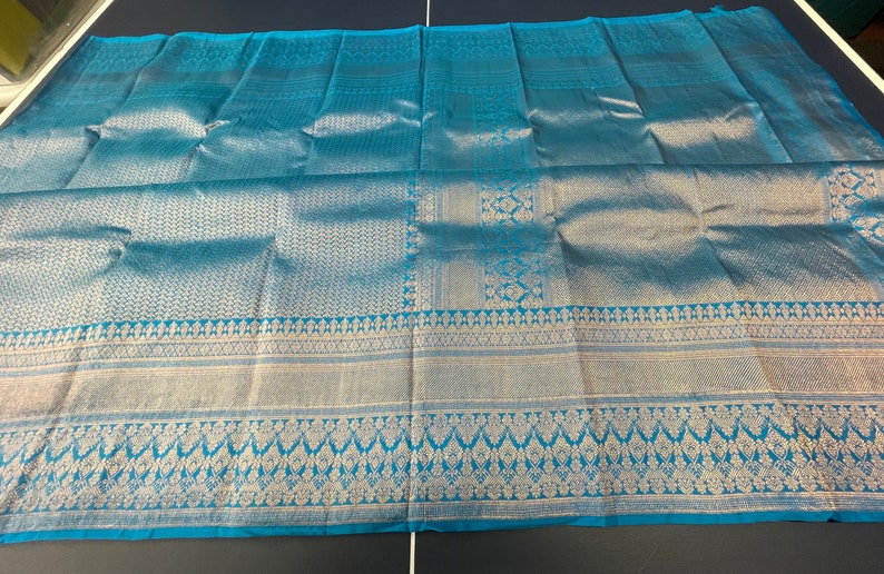 May include: A turquoise textile with intricate silver patterns. The fabric features detailed geometric and floral designs along the borders and central sections. The overall appearance is luxurious and elegant, suitable for apparel or home decor.