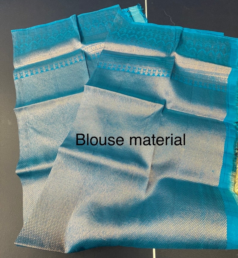 May include: Teal blouse material with a textured weave and decorative border. The fabric is folded, displaying the intricate pattern and the text "Blouse material". The material has a sheen and a soft appearance.