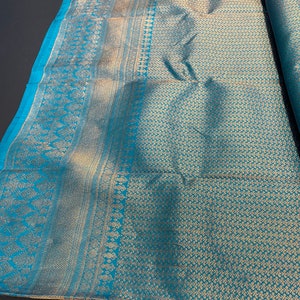 May include: A folded textile with a teal and gold pattern. The fabric has a detailed design along the edges and a repeating floral pattern across the main body. The teal color is a vibrant contrast to the gold, creating a luxurious look.