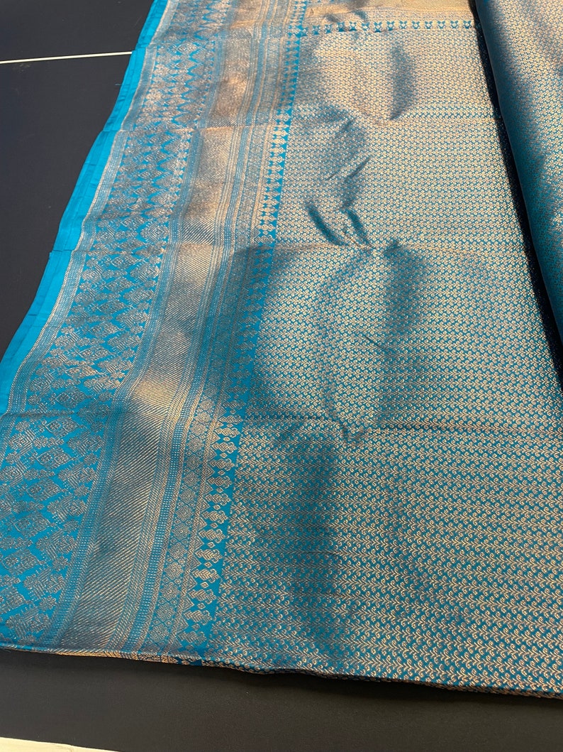 May include: A folded textile with a teal and gold pattern. The fabric has a detailed design along the edges and a repeating floral pattern across the main body. The teal color is a vibrant contrast to the gold, creating a luxurious look.