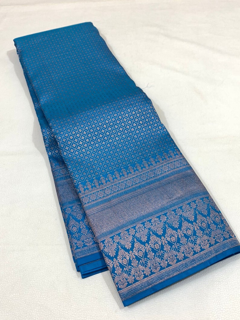 May include: A folded teal textile with a detailed pattern. The fabric has a repeating geometric design across the main body and a decorative border with floral and geometric elements. The textile is likely used for clothing or home decor.