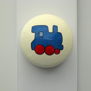 May include: A circular cabinet knob in cream, featuring a blue train illustration with red wheels. The knob is mounted on a white surface, suitable for a child's bedroom or nursery.