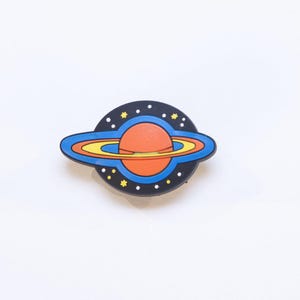 May include: A planet-shaped badge featuring a red planet, yellow and blue rings, and a black background with white and yellow stars. This space-themed accessory is ideal for adding a touch of cosmic flair to any outfit.