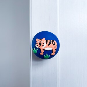 May include: A blue round knob with a tiger and green leaves design. The knob is made of plastic and has a smooth surface.