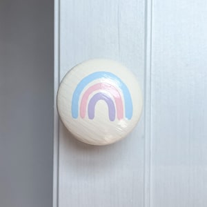 May include: A circular, cream-coloured cabinet knob featuring a pastel rainbow design in blue, pink, and purple. The knob is attached to a white cabinet door, providing a decorative element.