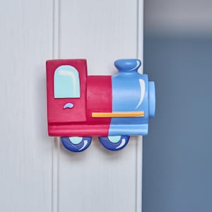 May include: A red and blue toy train door knob with a smiling face. The train has a yellow stripe and black wheels.