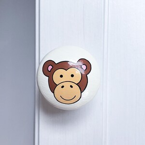 May include: A white ceramic door knob with a painted brown monkey face. The monkey has pink ears and a yellow smile.