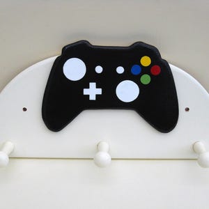 May include: White wooden wall-mounted coat rack shaped like a half-circle with a black painted video game controller in the centre. The controller has white buttons and colourful face buttons. There are three white hooks on the bottom.